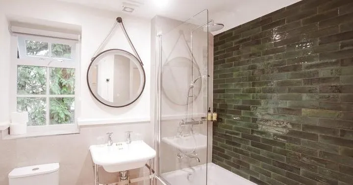 Bright modern bathroom with a white bathtub, glass shower screen, green tiled feature wall and round hanging mirror.