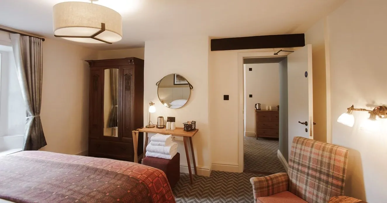 Warmly lit hotel bedroom with a wooden wardrobe, desk, plaid armchair and patterned bedspread.