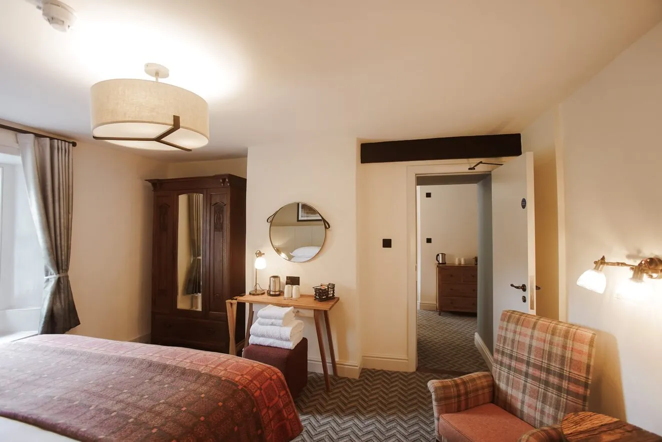 Warmly lit hotel bedroom with a wooden wardrobe, desk, plaid armchair and patterned bedspread.