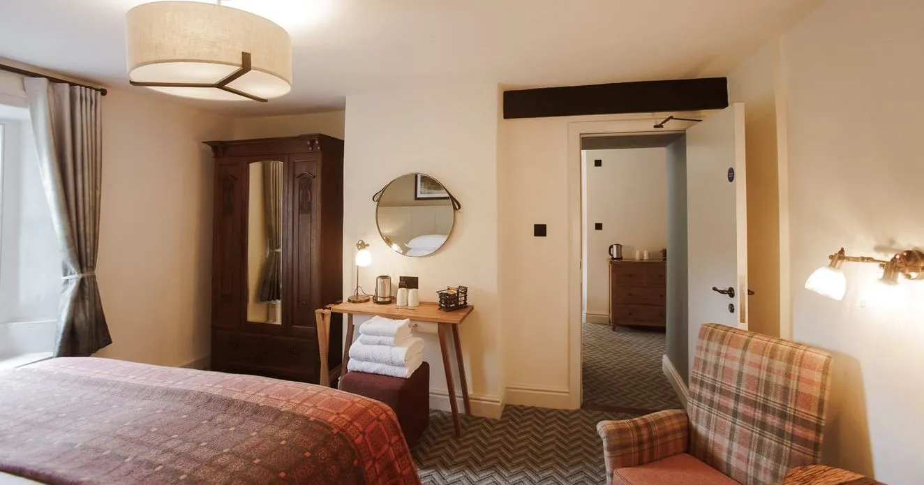 Warmly lit hotel bedroom with a wooden wardrobe, desk, plaid armchair and patterned bedspread.