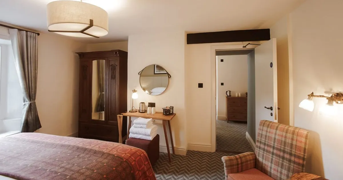 Warmly lit hotel bedroom with a wooden wardrobe, desk, plaid armchair and patterned bedspread.