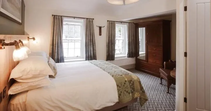 Warmly lit bedroom with a double bed, patterned throw, dark wood wardrobe and curtained windows.