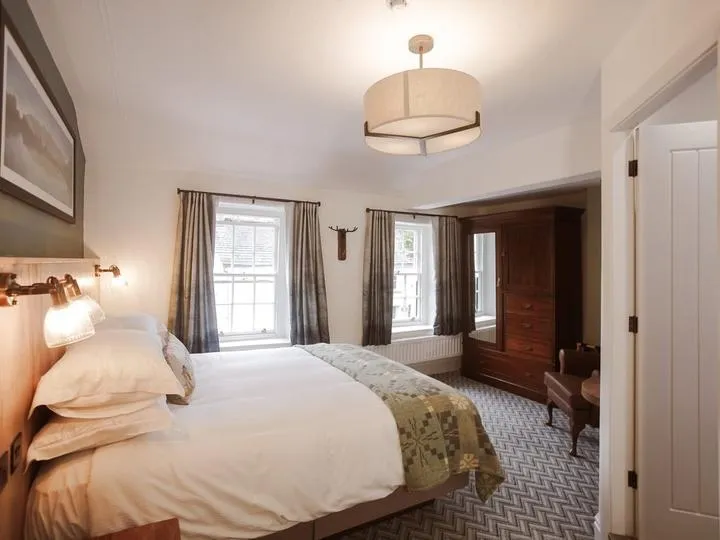 Warmly lit bedroom with a double bed, patterned throw, dark wood wardrobe and curtained windows.