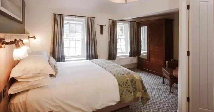 Warmly lit bedroom with a double bed, patterned throw, dark wood wardrobe and curtained windows.