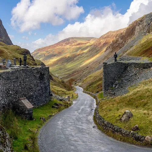 Winding road through a rugged Welsh mountain valley beside a historic slate cemetery under a blue sky.
