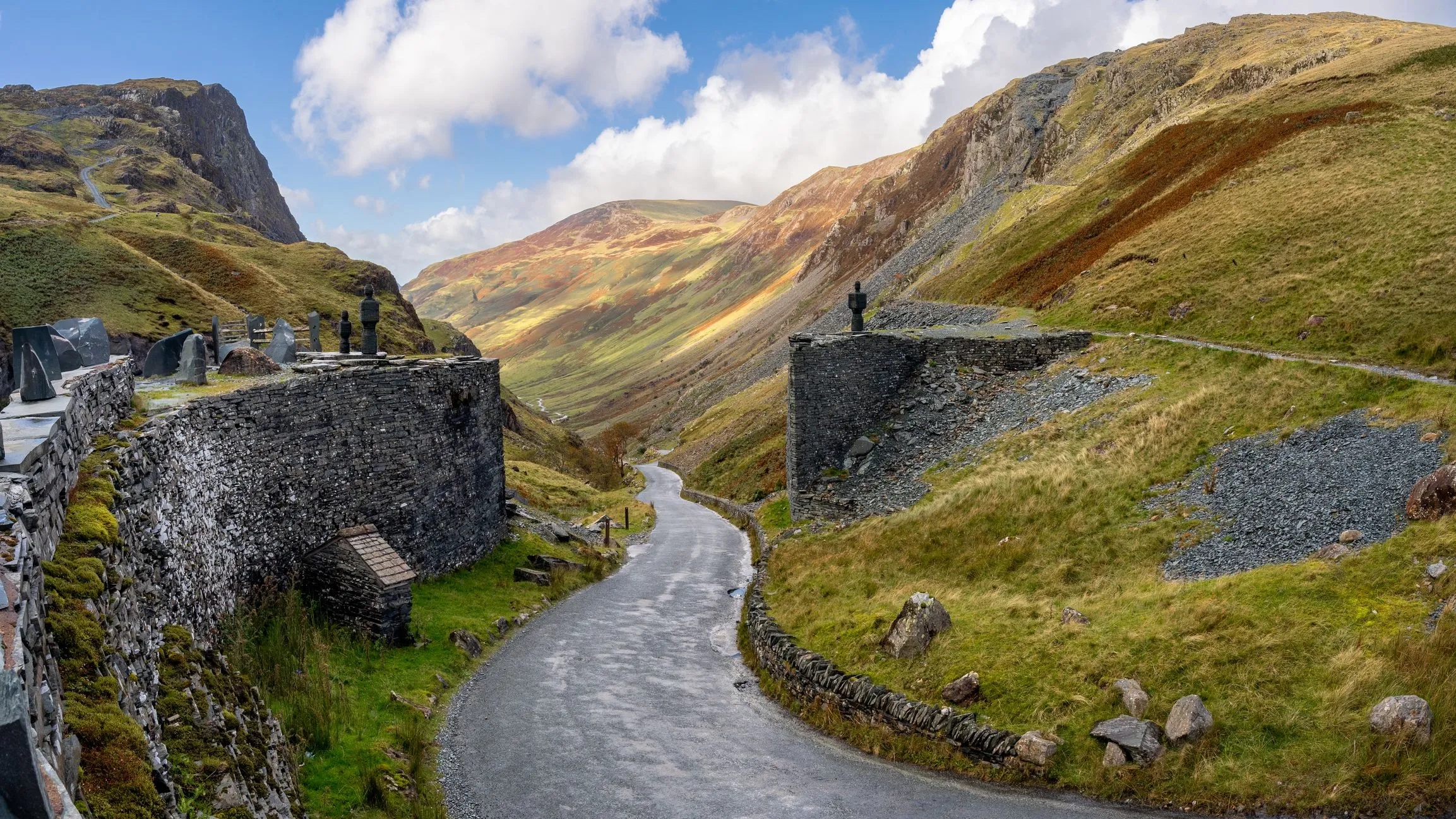 Winding road through a rugged Welsh mountain valley beside a historic slate cemetery under a blue sky.