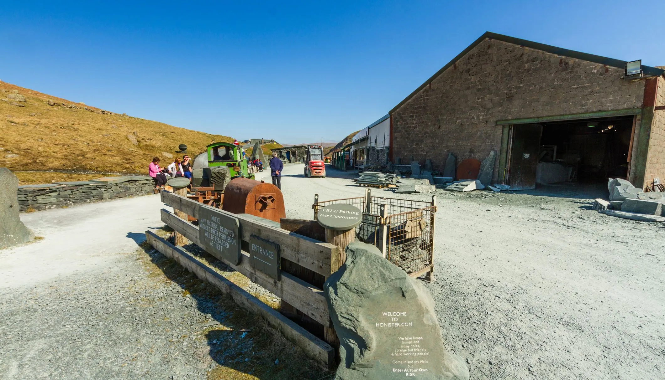 Visitors at the entrance to Honister Slate Mine, with stone workshop buildings and a green mine train beneath a clear blue sky.
