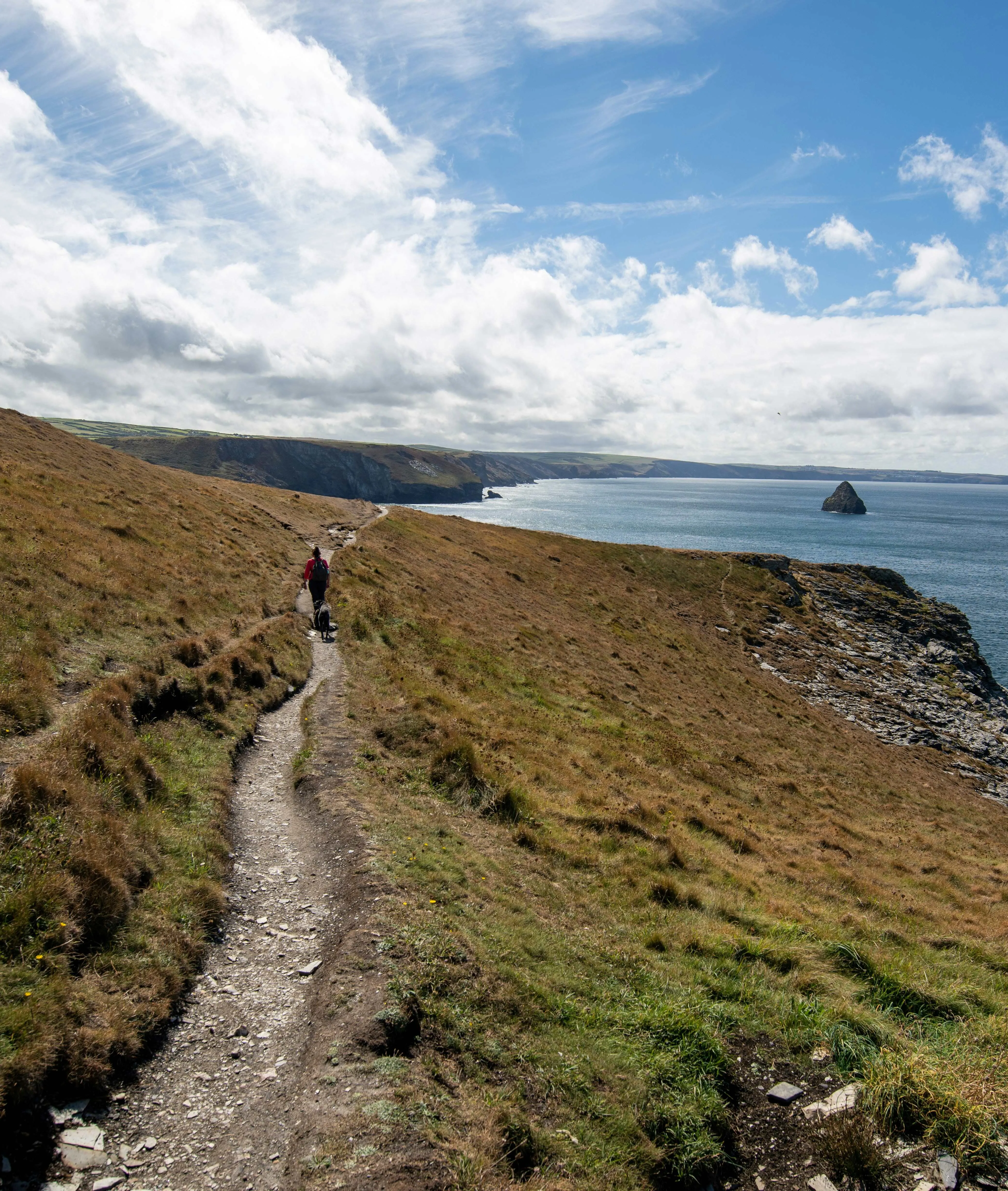 Hiker walking along a coastal cliff path above the sea under a partly cloudy blue sky