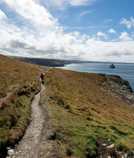 Hiker walking along a coastal cliff path above the sea under a partly cloudy blue sky