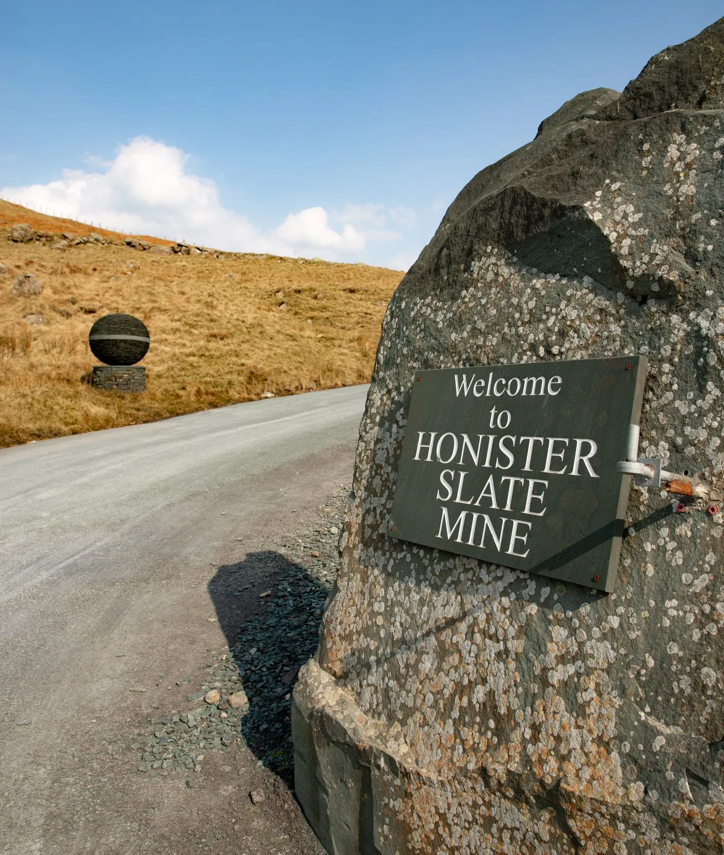 Welcome sign for Honister Slate Mine mounted on a large boulder beside a rural road