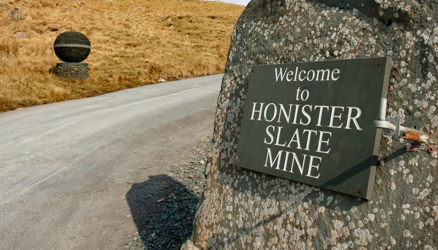 Welcome sign for Honister Slate Mine mounted on a large boulder beside a rural road