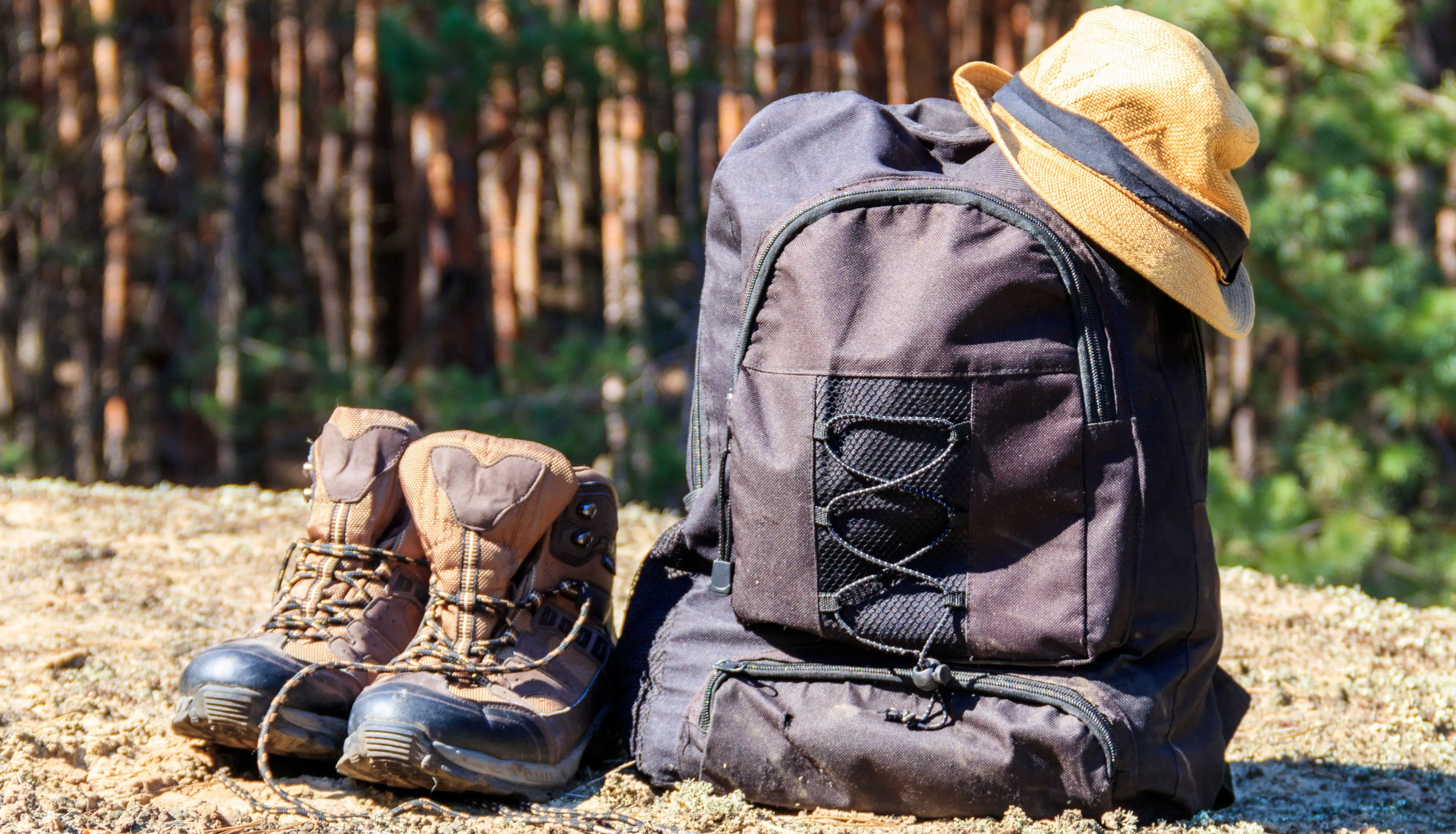 Hiking boots beside a black rucksack with a straw hat in a sunlit forest