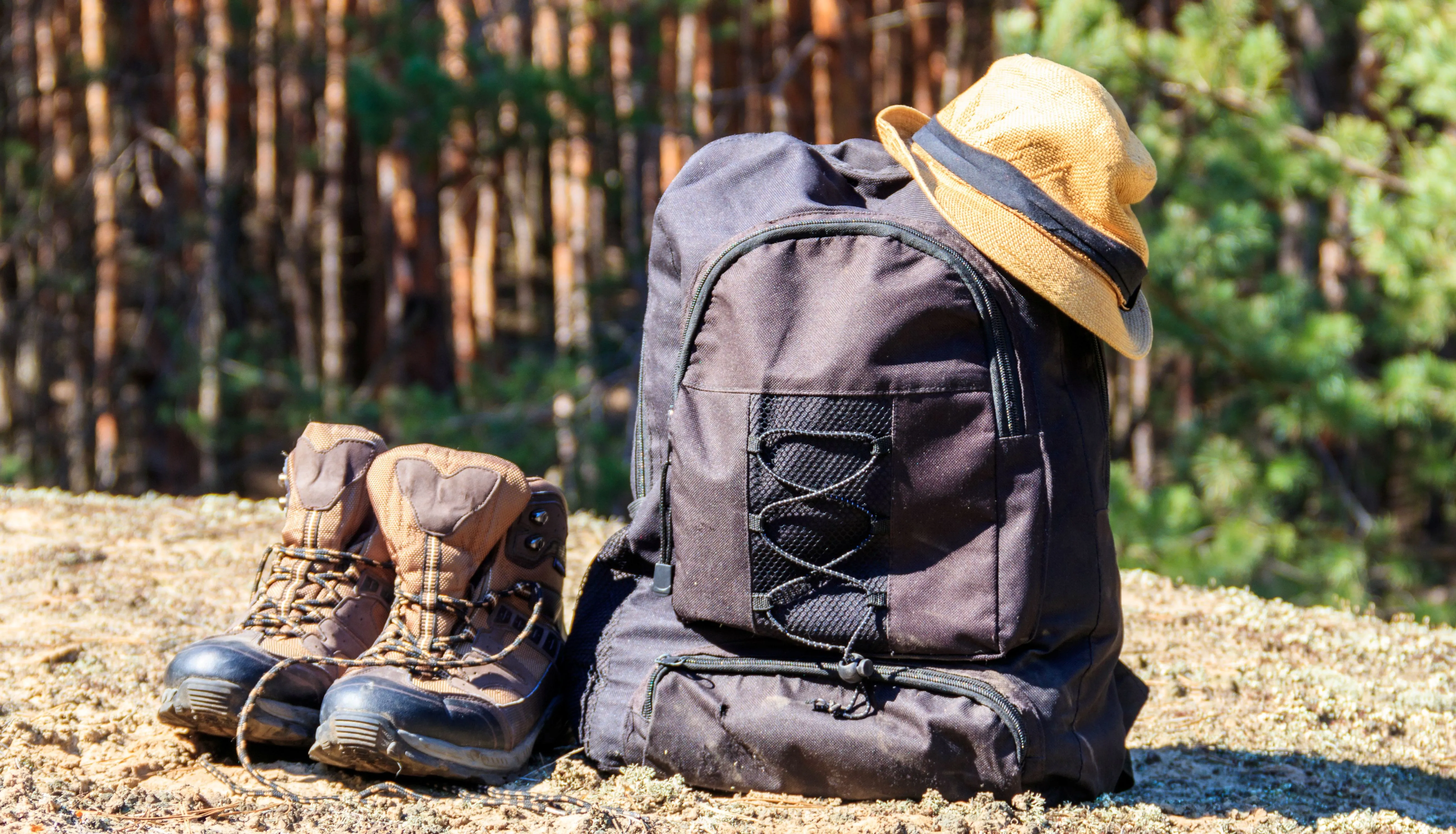 Hiking boots beside a black rucksack with a straw hat in a sunlit forest