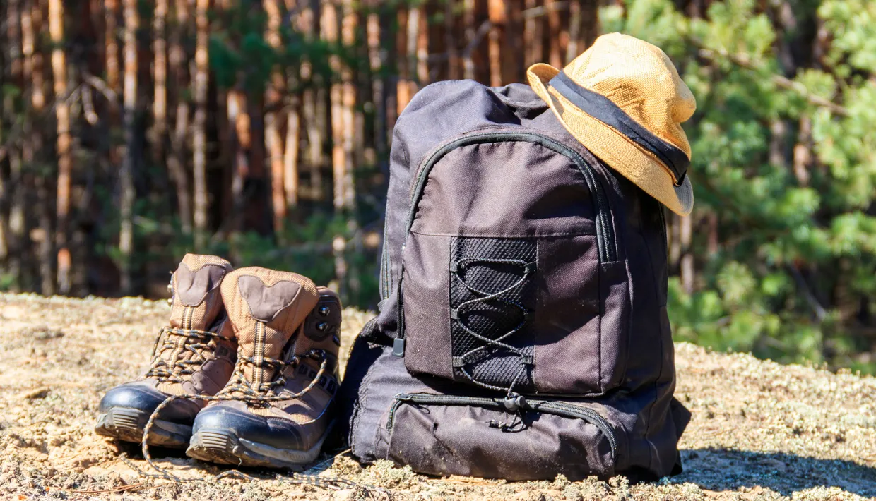Hiking boots beside a black rucksack with a straw hat in a sunlit forest