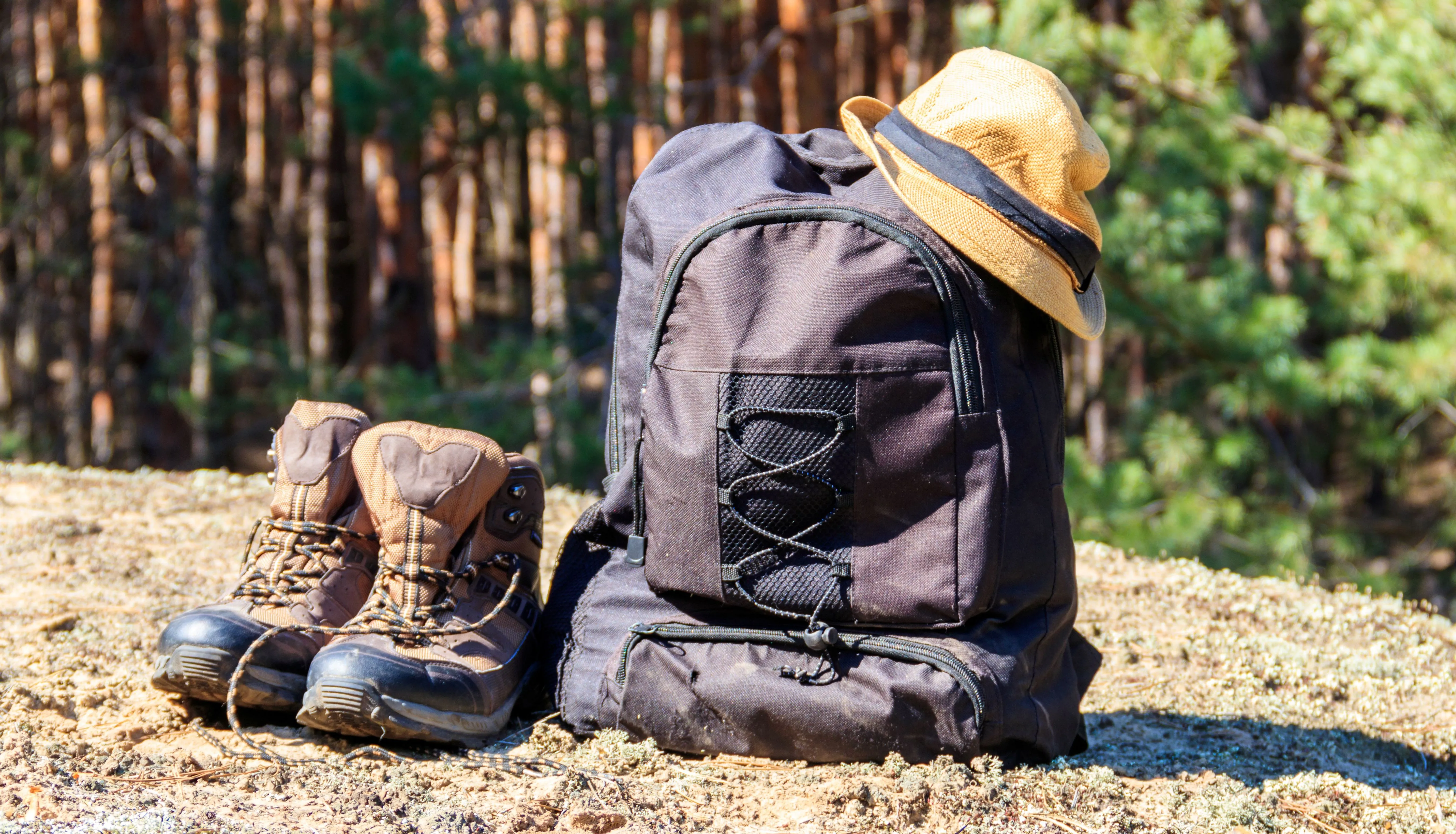 Hiking boots beside a black rucksack with a straw hat in a sunlit forest