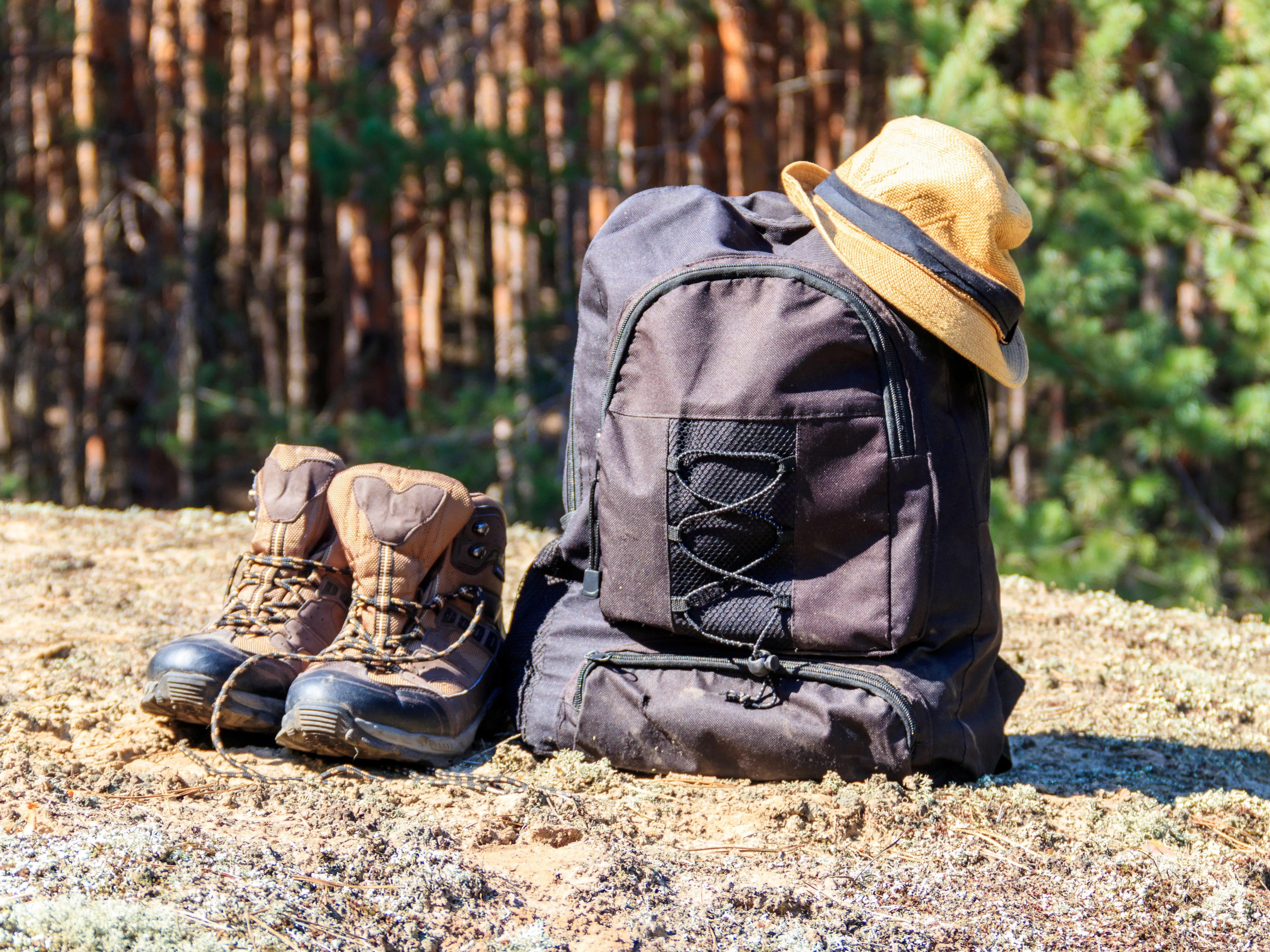 Hiking boots beside a black rucksack with a straw hat in a sunlit forest