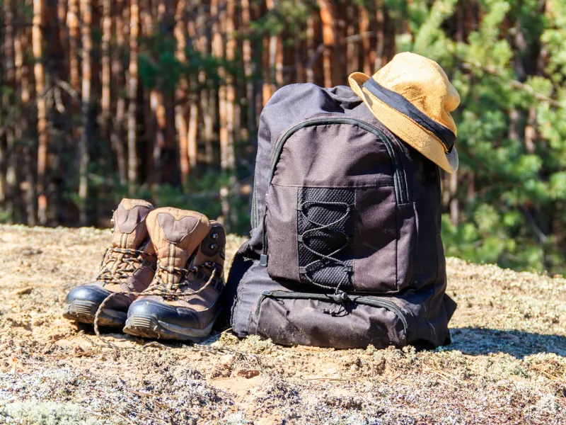 Hiking boots beside a black rucksack with a straw hat in a sunlit forest