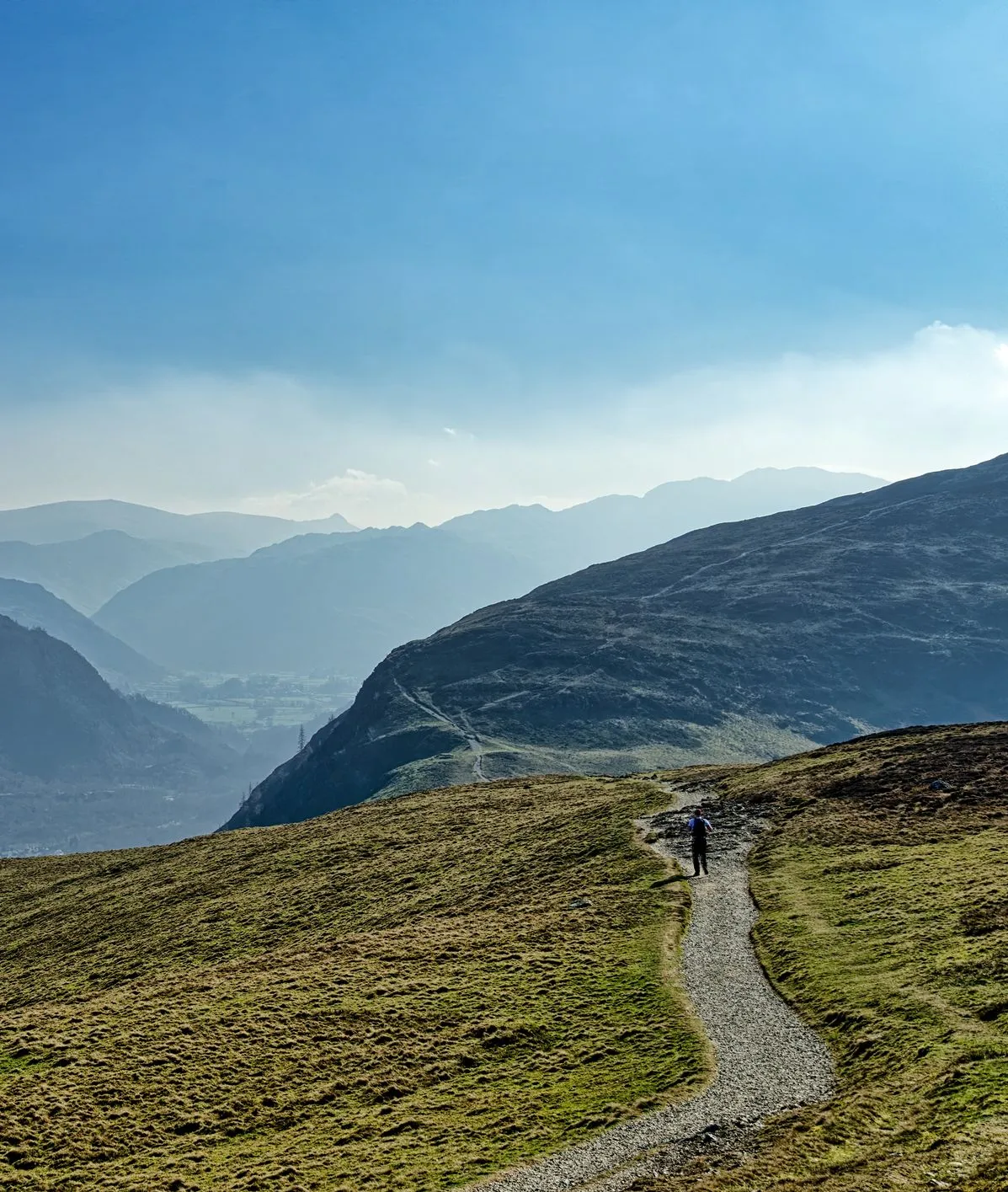 Lone hiker walking along a mountain path above a misty valley under a blue sky
