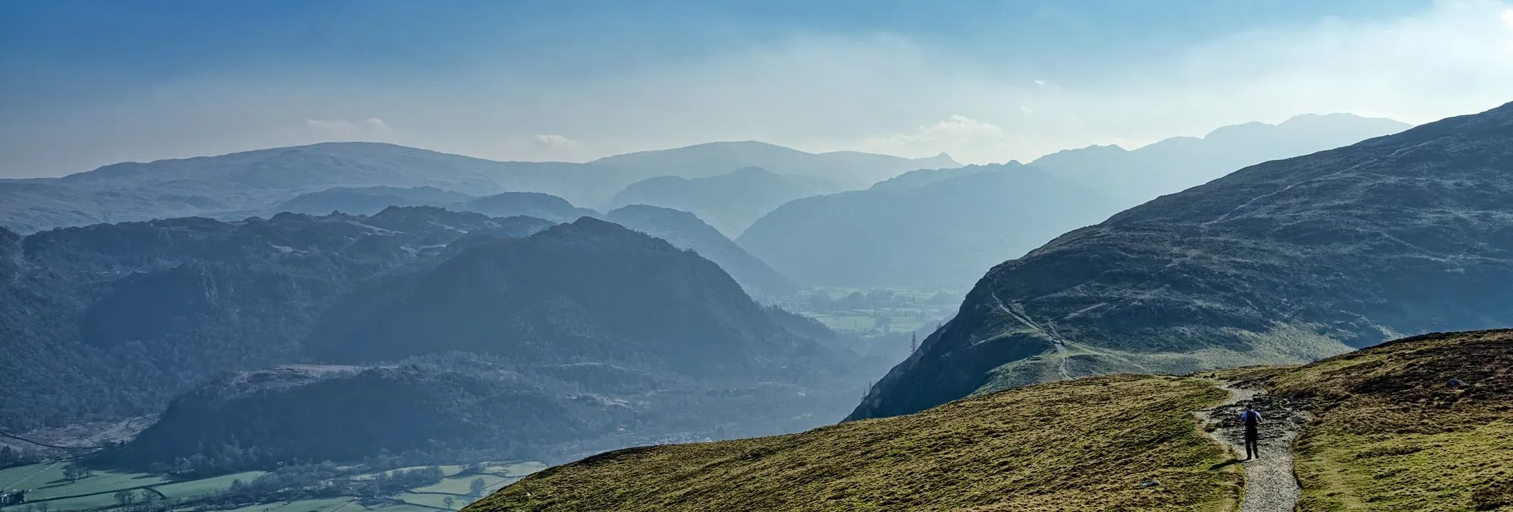 Lone hiker walking along a mountain path above a misty valley under a blue sky