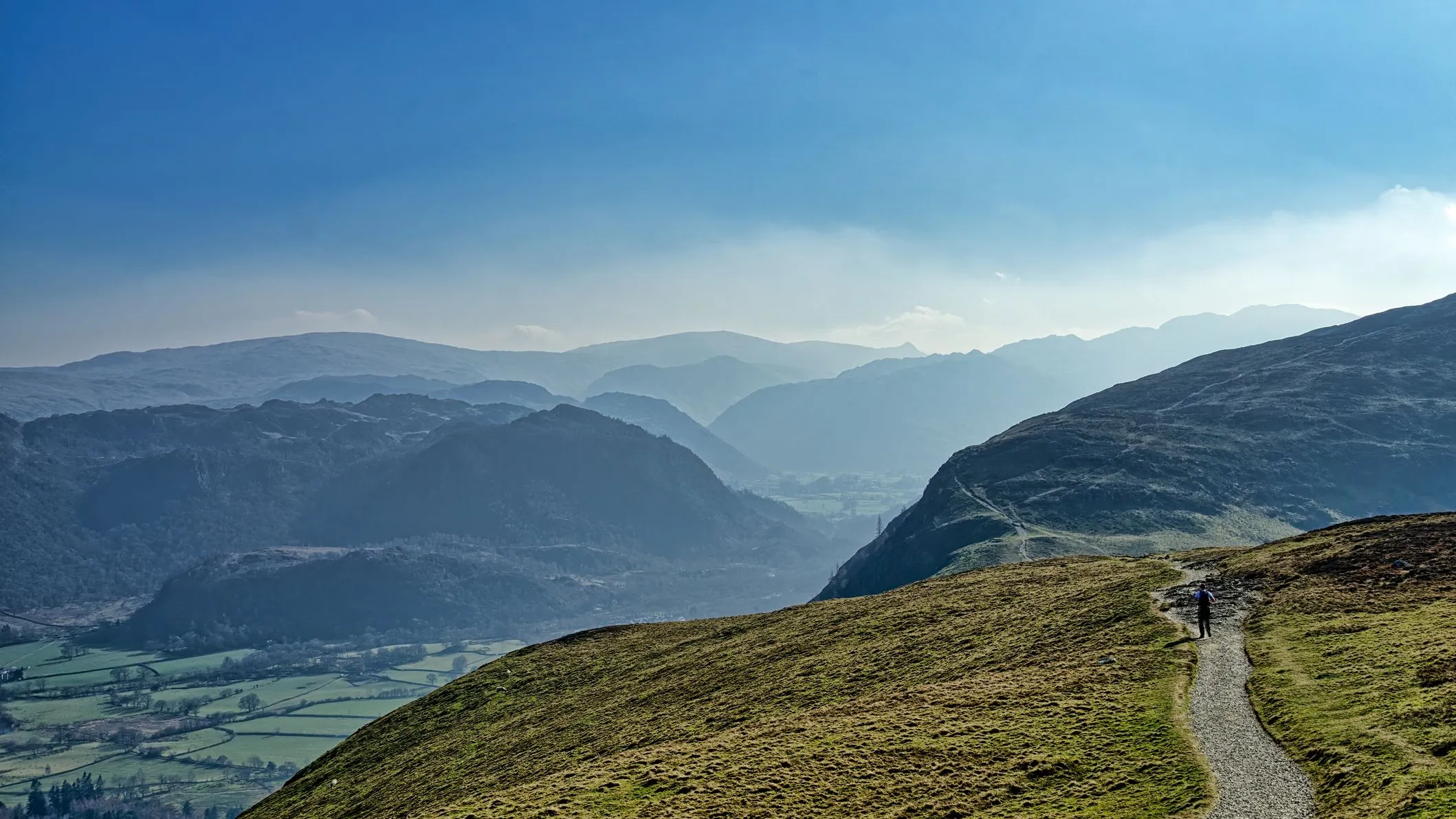 Lone hiker walking along a mountain path above a misty valley under a blue sky