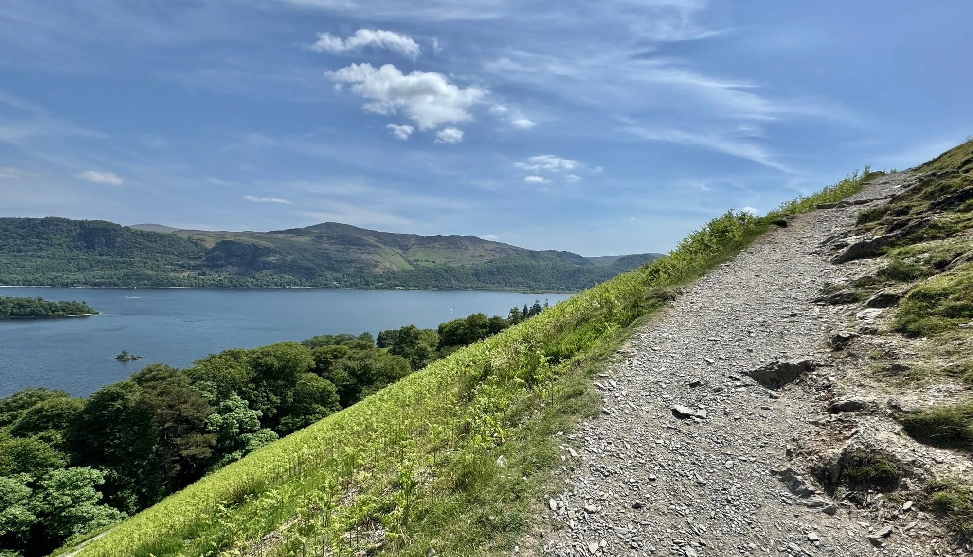 Rocky hillside path overlooking a blue lake, wooded shoreline and green mountains beneath a bright cloudy sky.