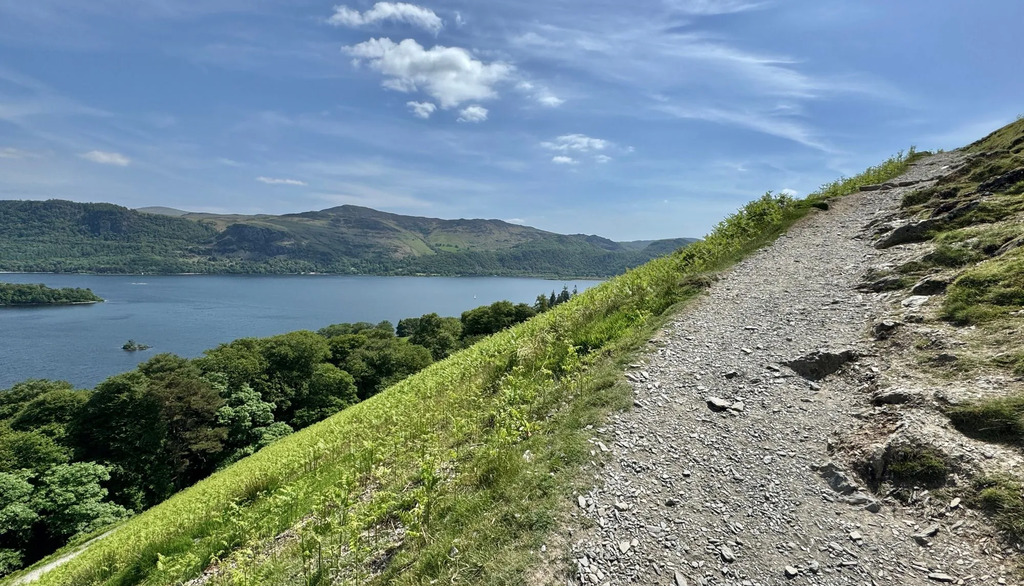 Rocky hillside path overlooking a blue lake, wooded shoreline and green mountains beneath a bright cloudy sky.