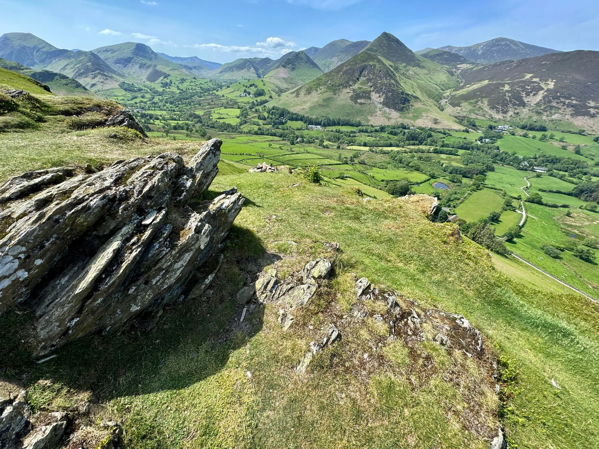 Rocky hillside overlooking a green valley and rugged mountains beneath a clear blue sky