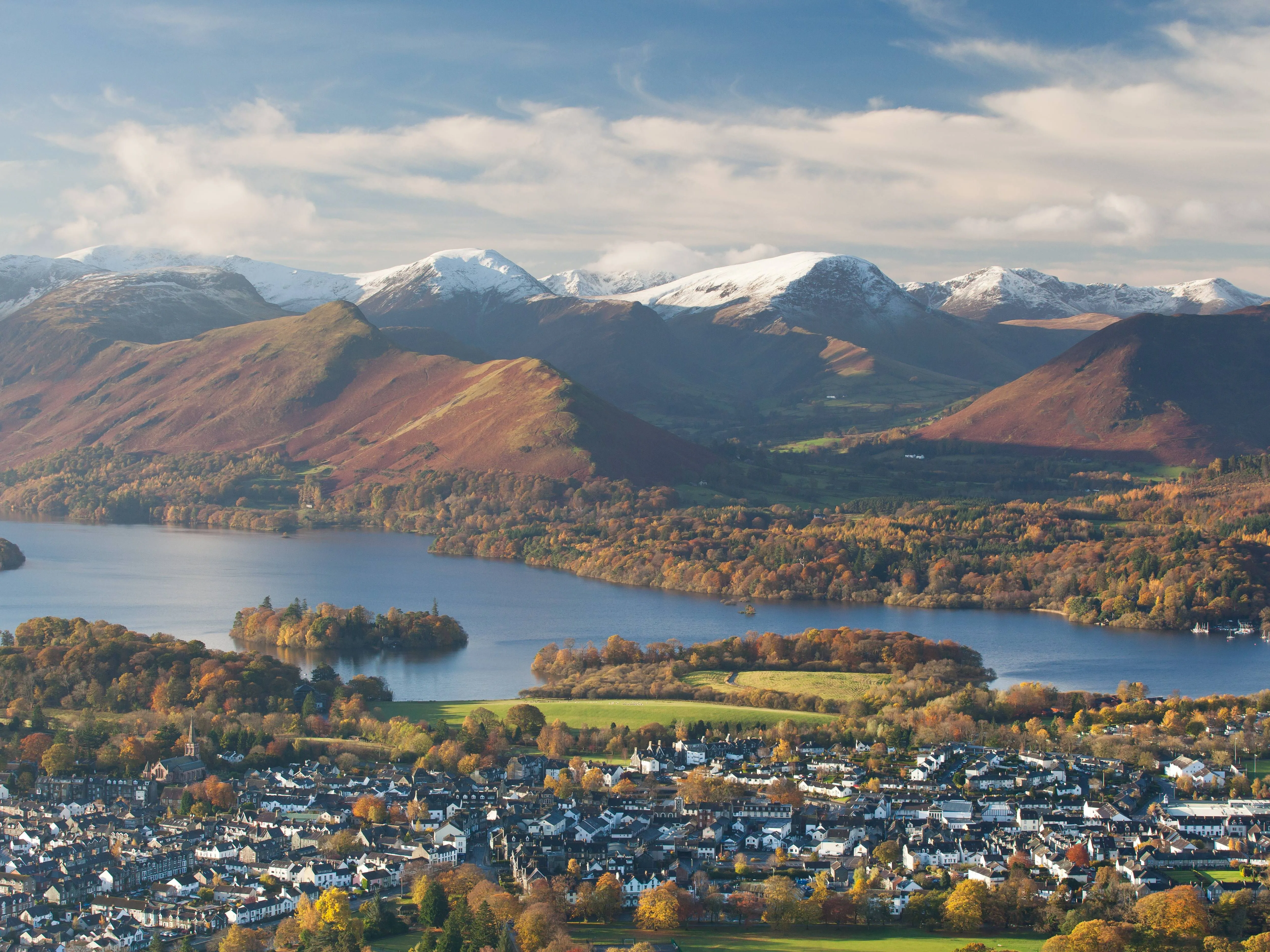 Aerial view of Keswick beside Derwentwater, with autumn woodland and snow-covered Lake District fells under a cloudy sky.