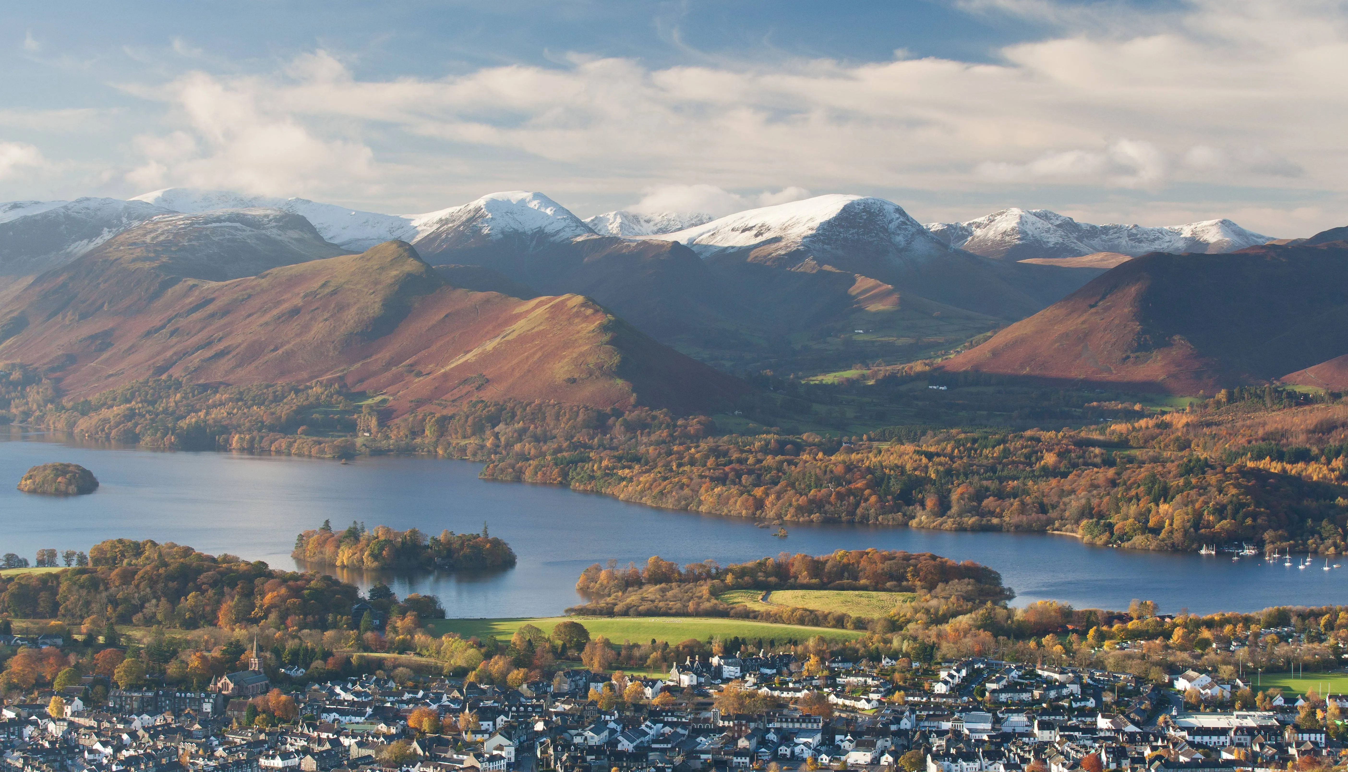 Aerial view of Keswick beside Derwentwater, with autumn woodland and snow-covered Lake District fells under a cloudy sky.