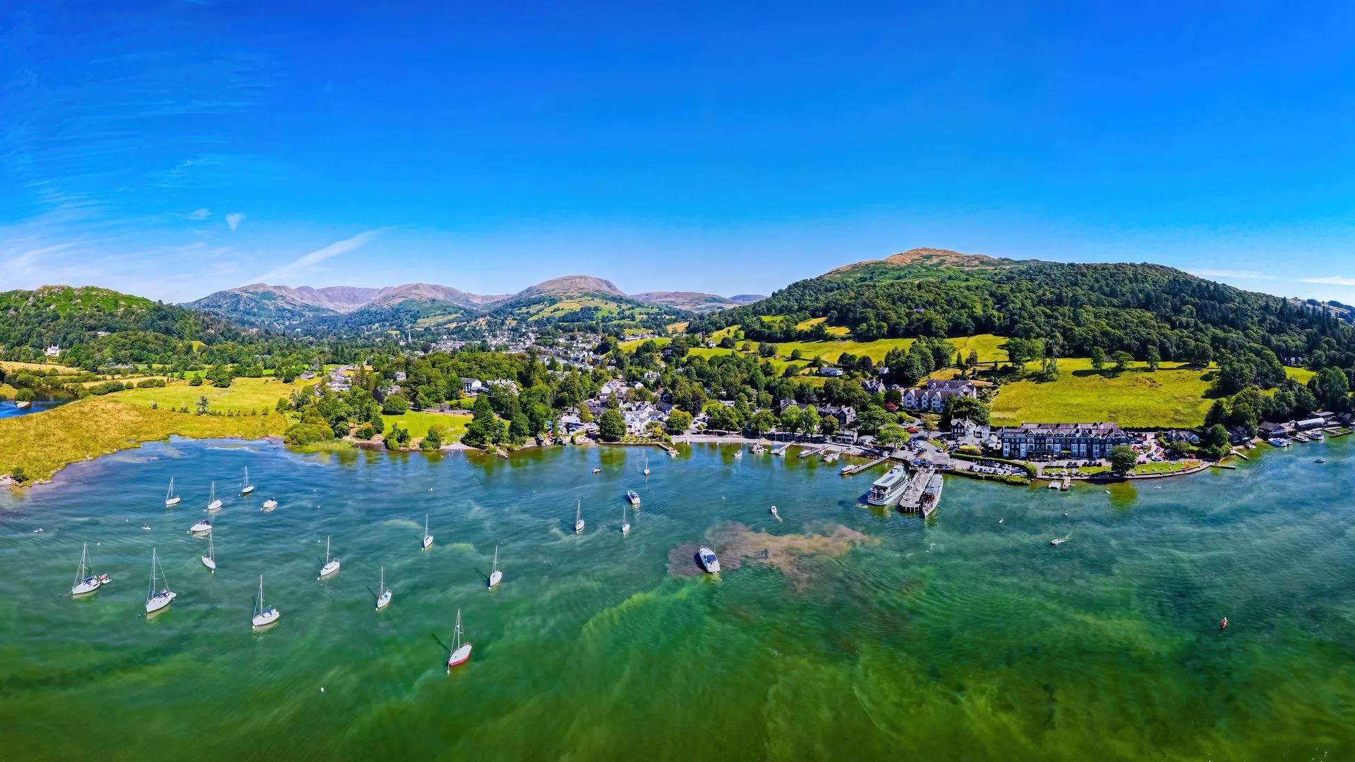 Aerial view of a lakeside village, green hills and anchored sailboats under a bright blue sky.