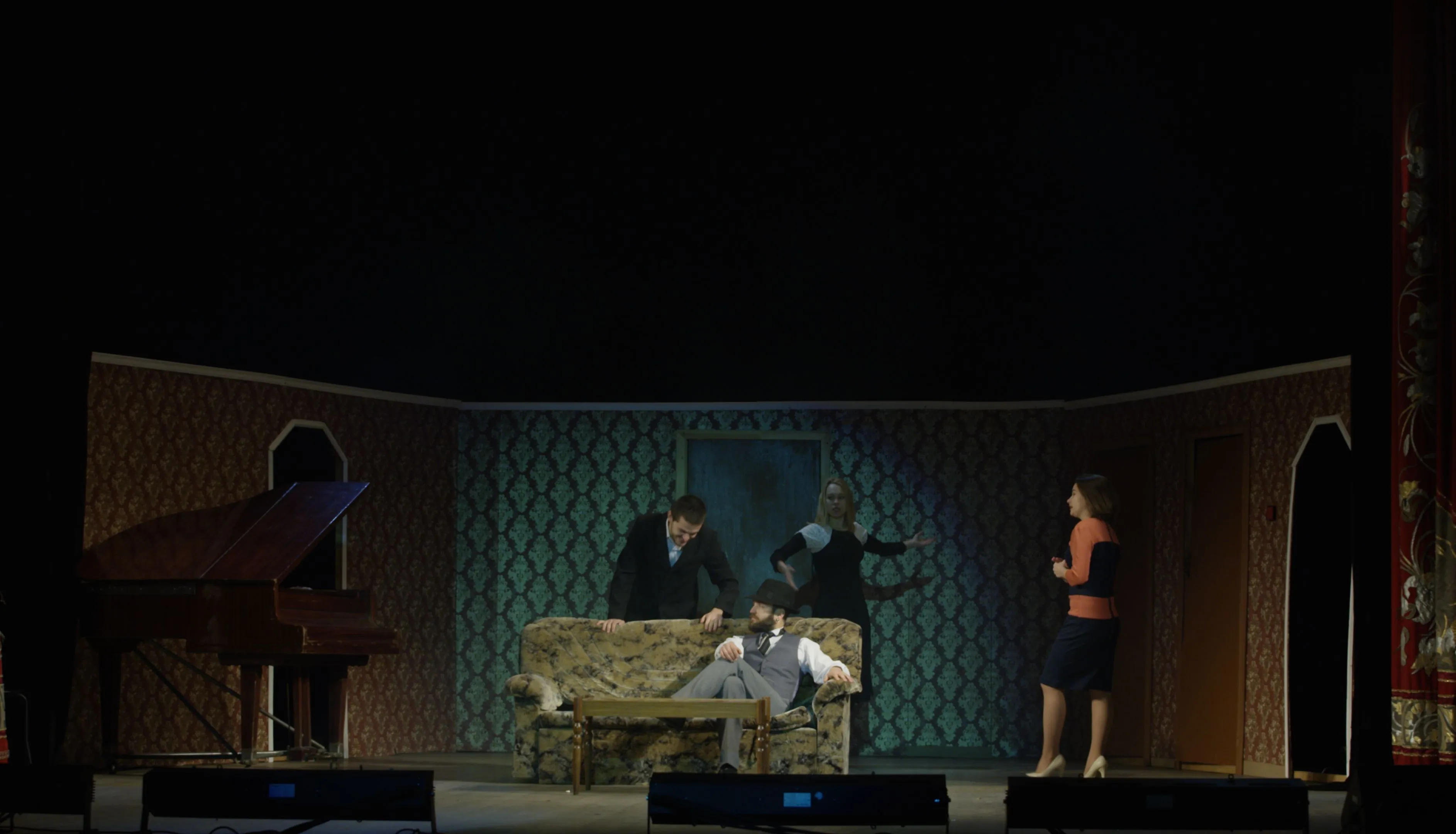 Theatre stage set with a grand piano, patterned sofa and four actors in a domestic drama scene.