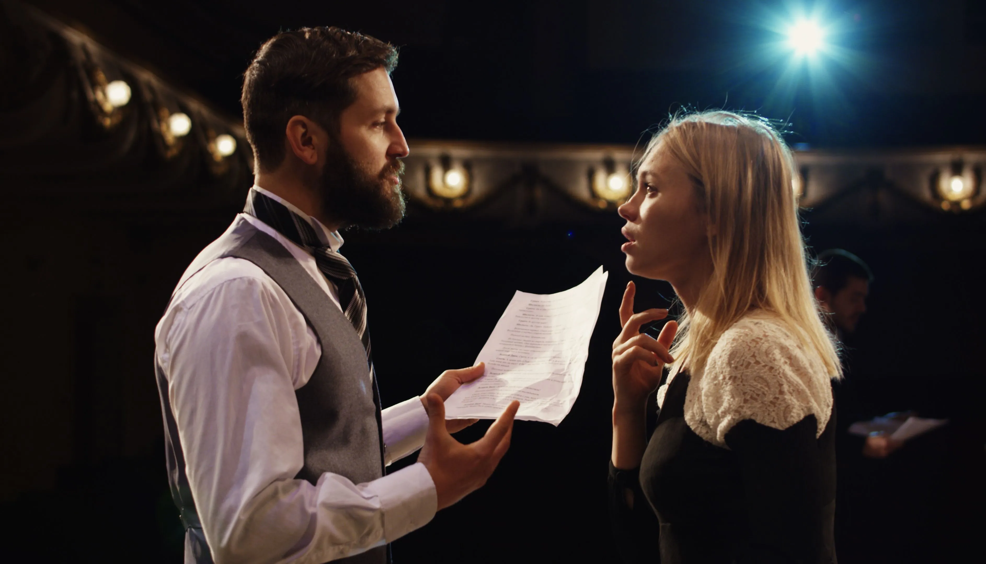 Male and female actors rehearsing lines from a script on a dimly lit theatre stage.