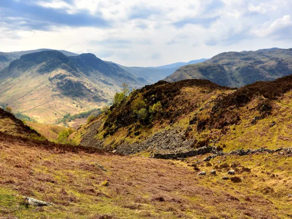 Sunlit mountain valley with rugged grassy hills, dry-stone walls and grazing sheep beneath a cloudy blue sky.
