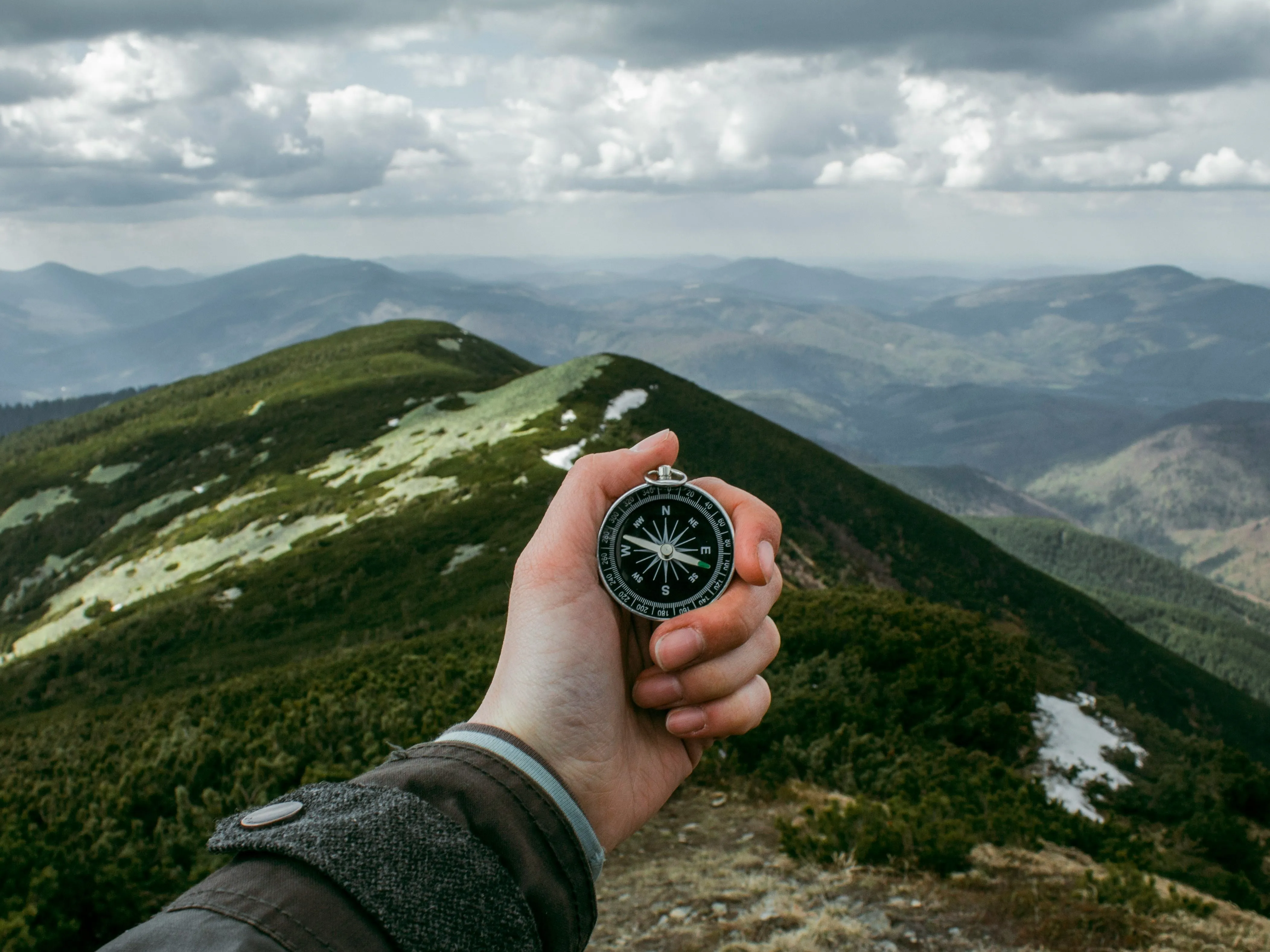 Hand holding a compass above a green mountain ridge beneath cloudy skies
