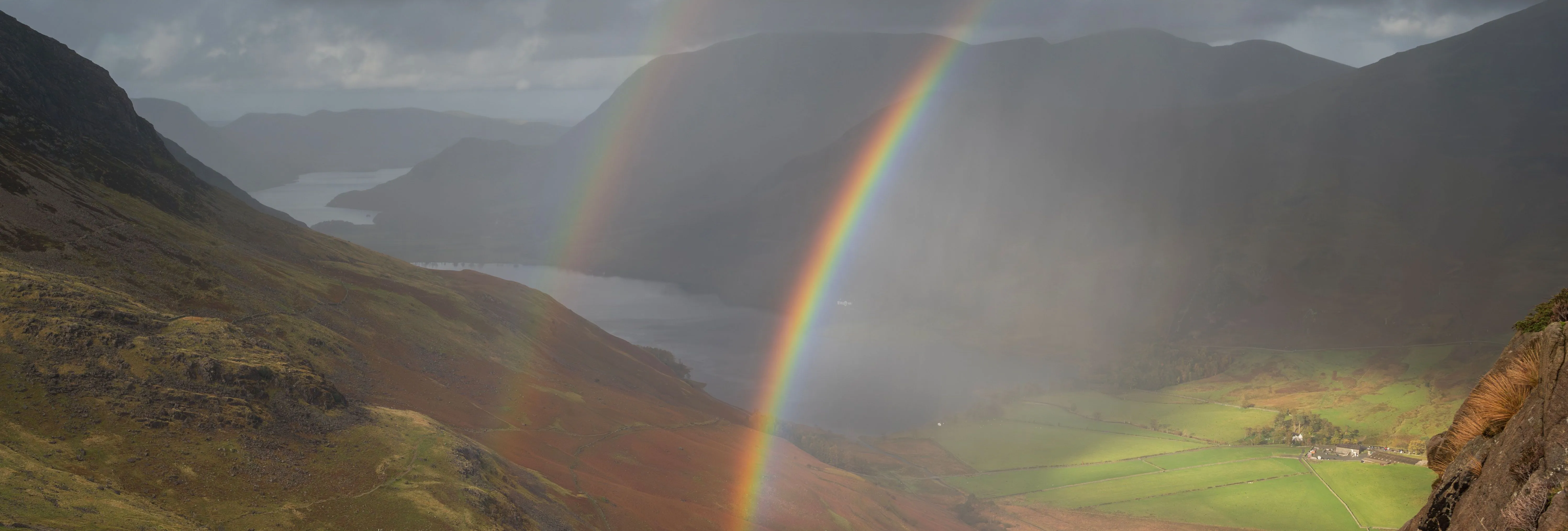 Vivid rainbow over a dramatic mountain valley with a lake, green fields and storm clouds.
