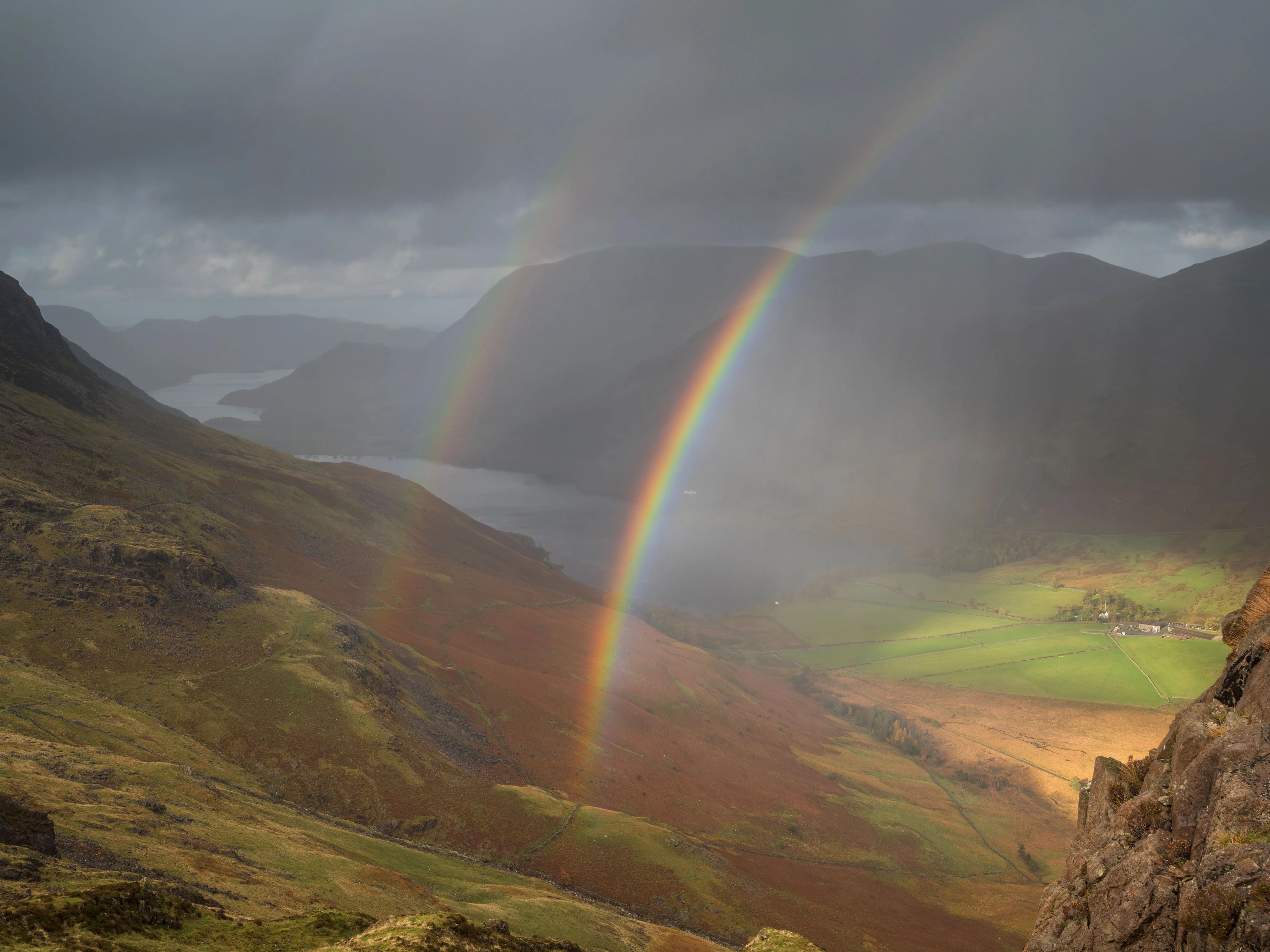 Vivid rainbow over a dramatic mountain valley with a lake, green fields and storm clouds.