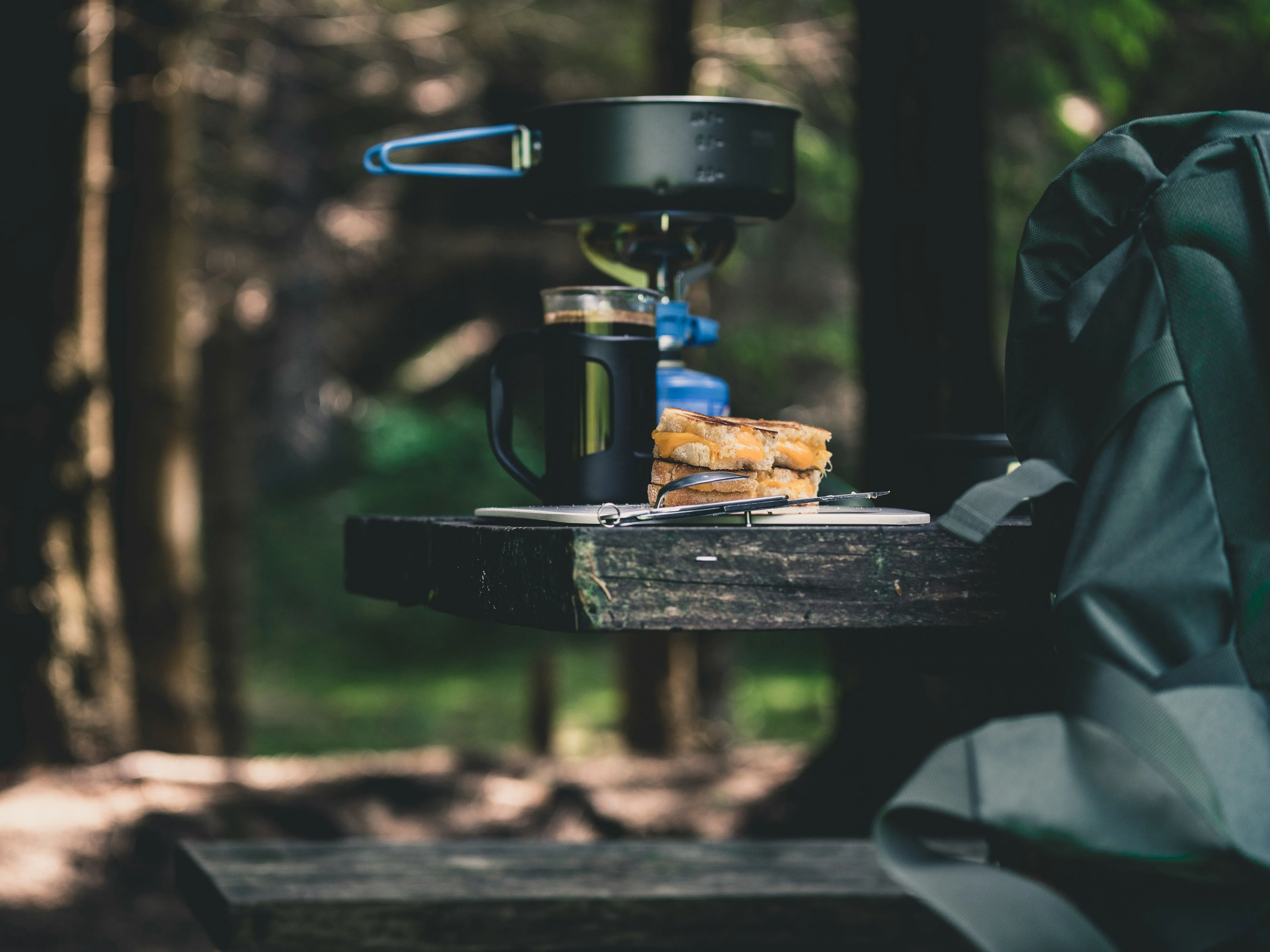 Camping stove, coffee mug and toasted sandwich on a wooden bench in a forest beside a backpack