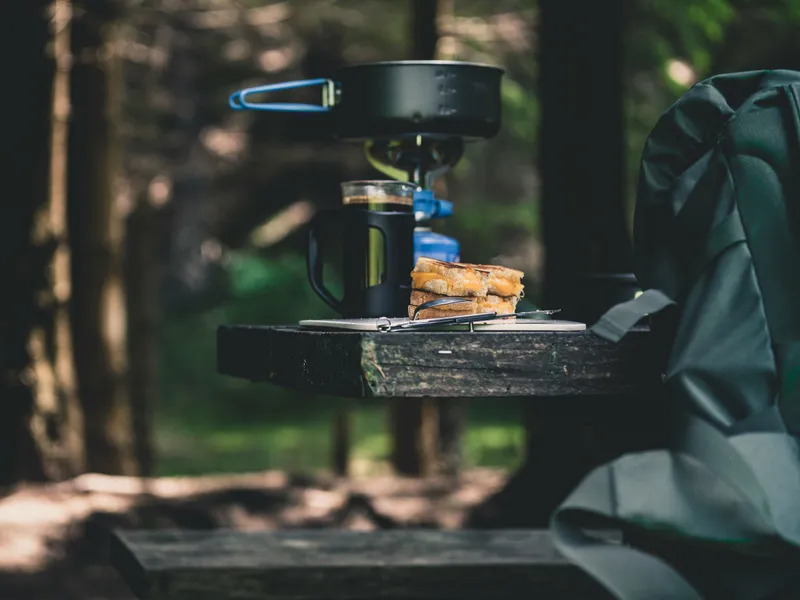Camping stove, coffee mug and toasted sandwich on a wooden bench in a forest beside a backpack