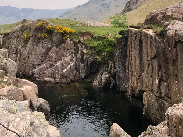 Rocky mountain gorge with a dark pool, grassy slopes and yellow wildflowers beneath a cloudy sky