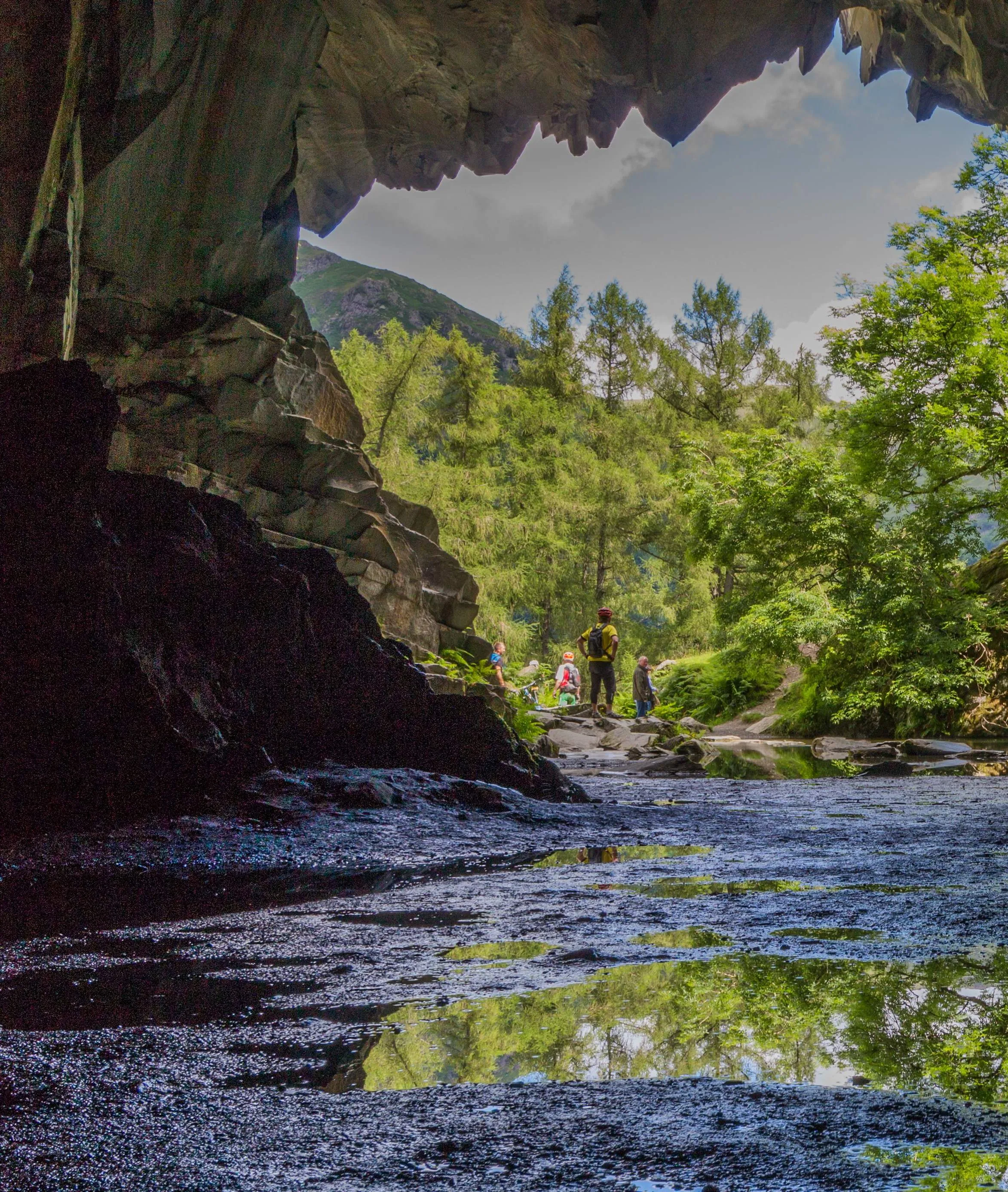 View from inside a rocky cave towards a sunlit tree, shallow water and distant hikers.