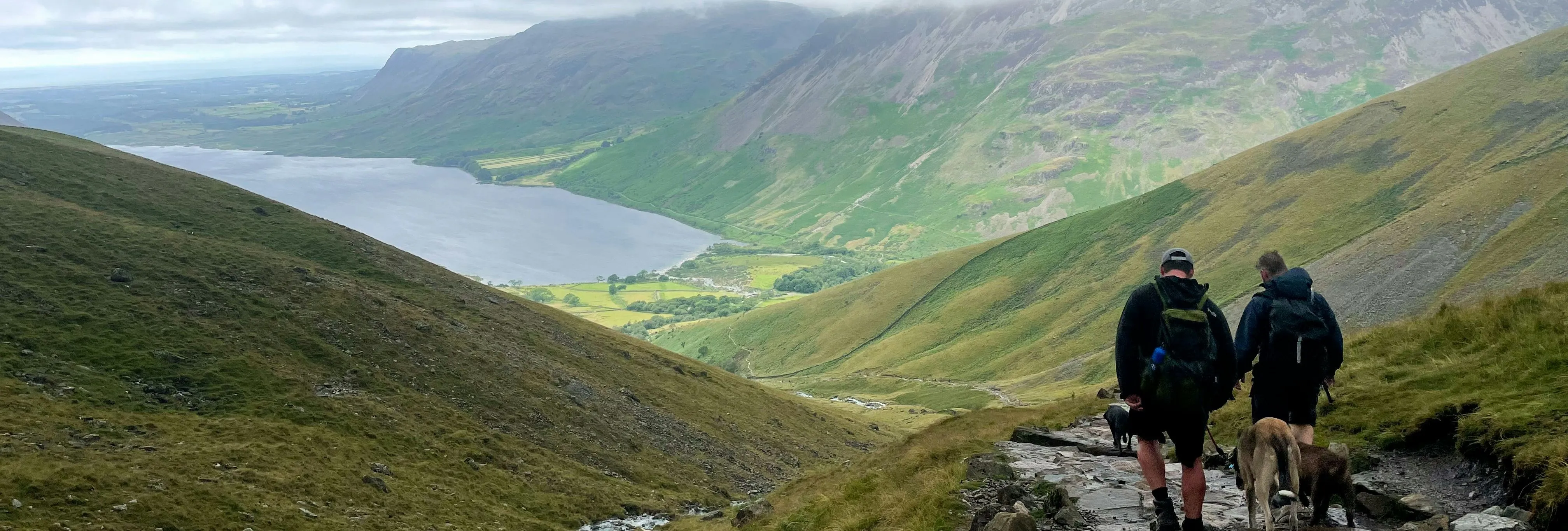 Two hikers with dogs descend a rocky mountain path overlooking a lake and green valley beneath low clouds.