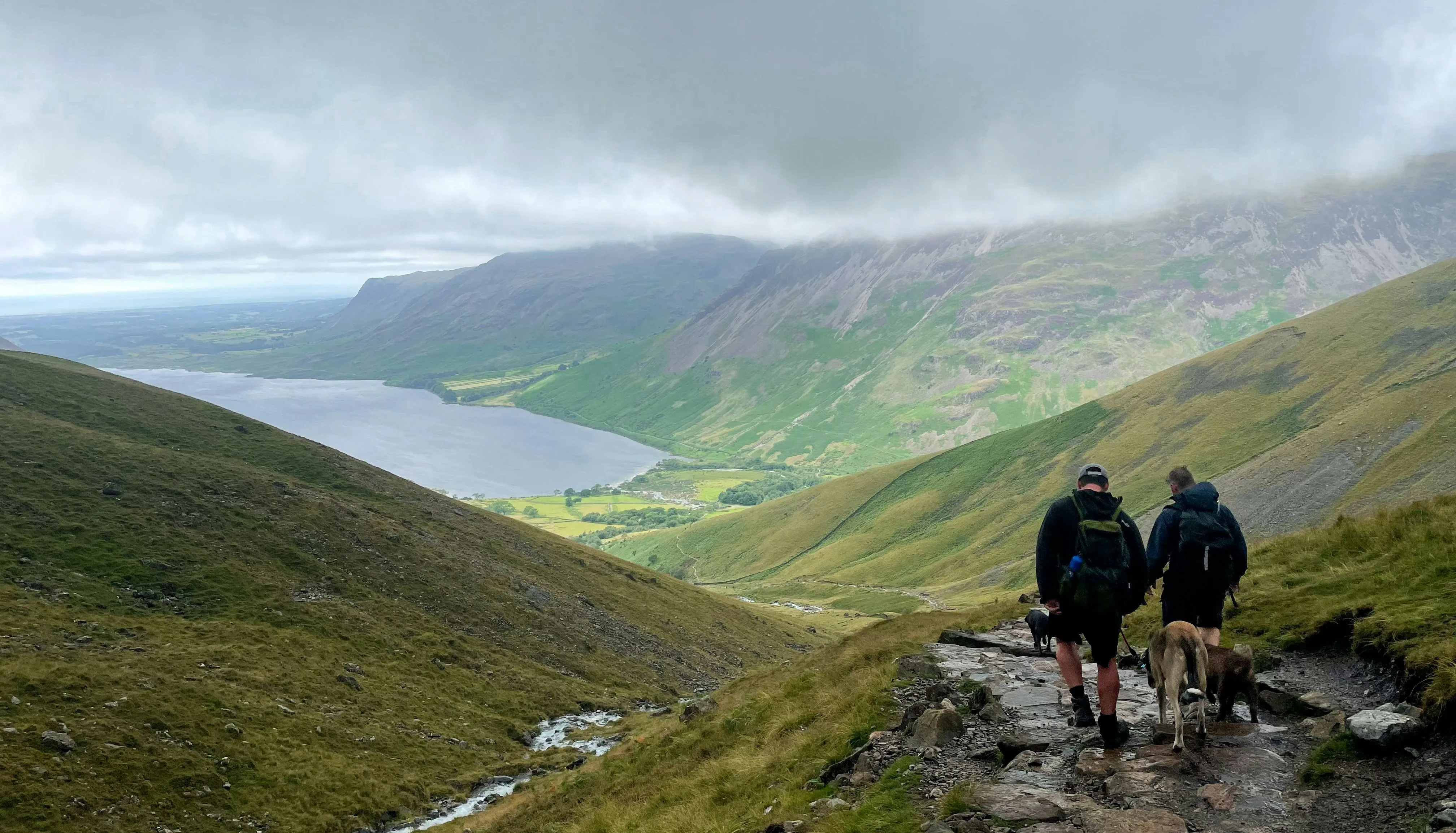 Two hikers with dogs descend a rocky mountain path overlooking a lake and green valley beneath low clouds.