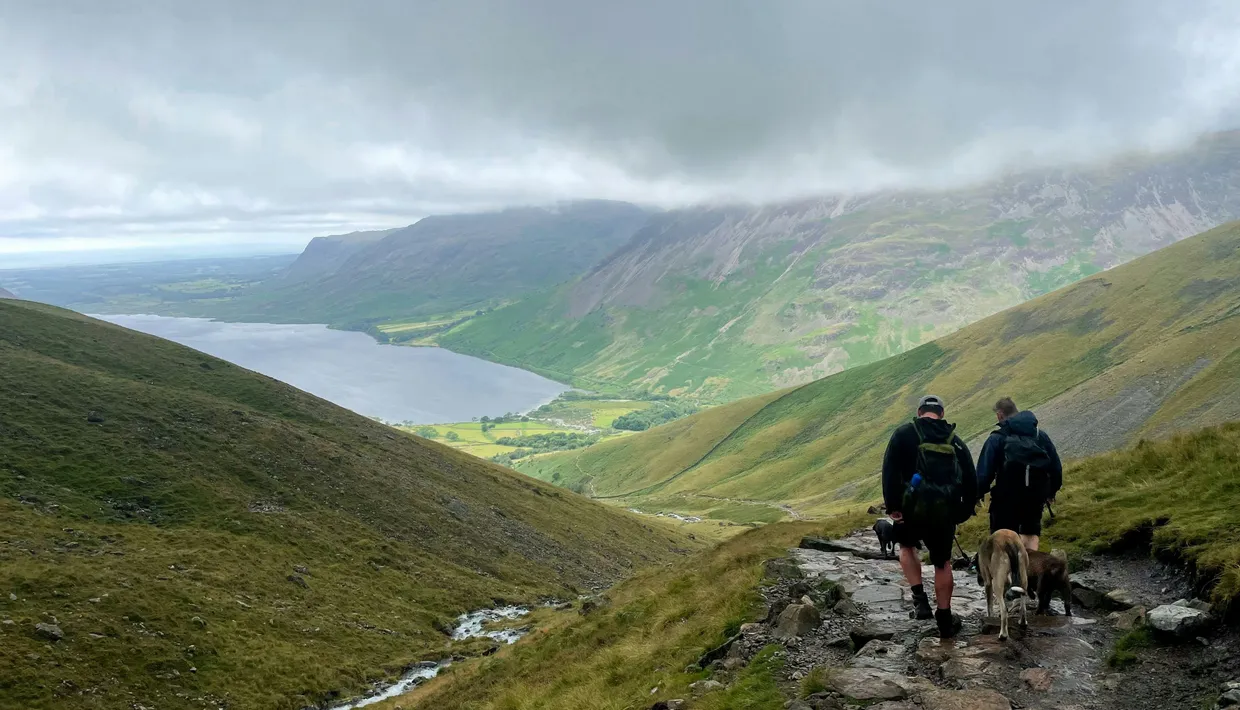 Two hikers with dogs descend a rocky mountain path overlooking a lake and green valley beneath low clouds.