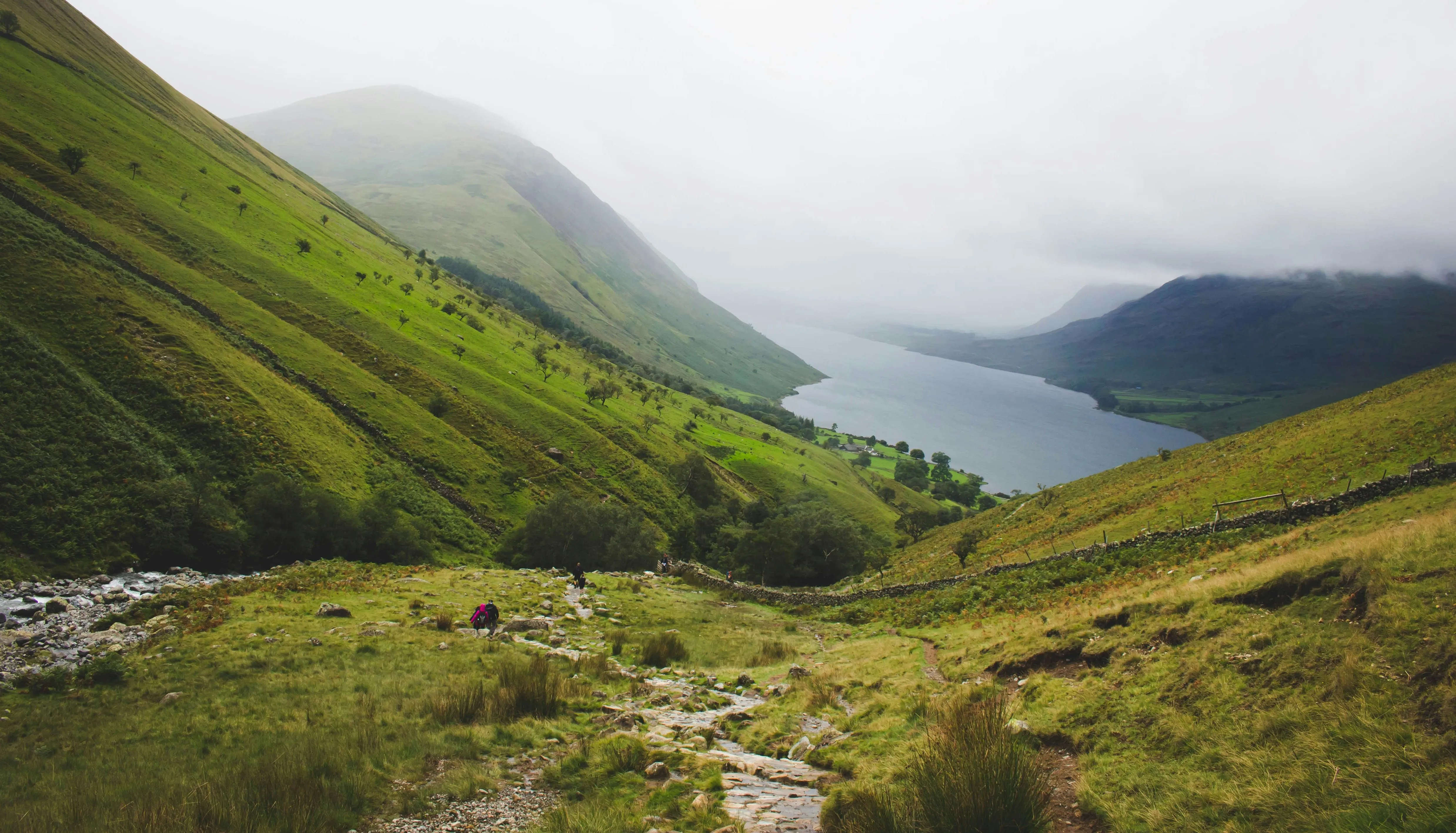 Misty green valley with a mountain lake, stone path and distant hikers