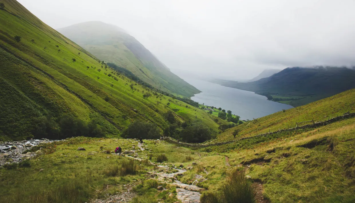 Misty green valley with a mountain lake, stone path and distant hikers