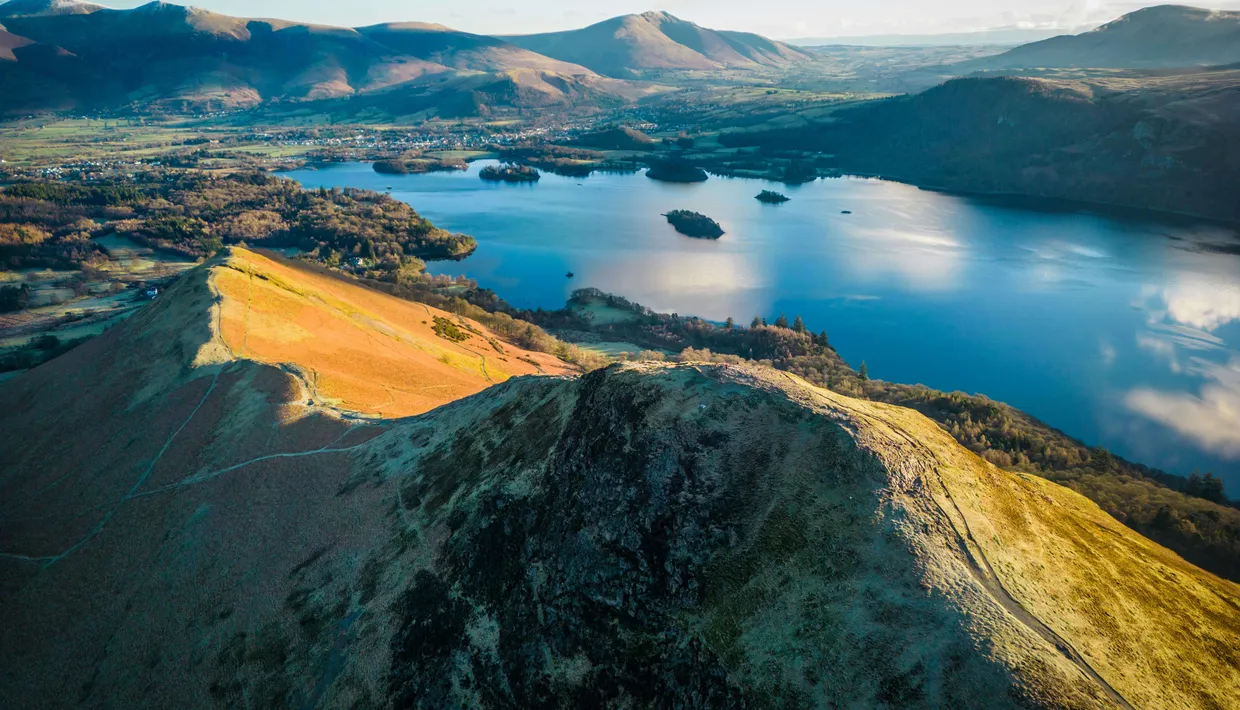 Aerial view of a sunlit mountain ridge overlooking a blue lake with wooded islands and surrounding hills.
