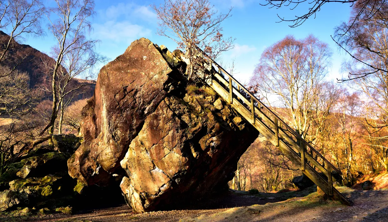 Wooden staircase climbing beside a large rocky outcrop in a sunlit woodland with bare birch trees.
