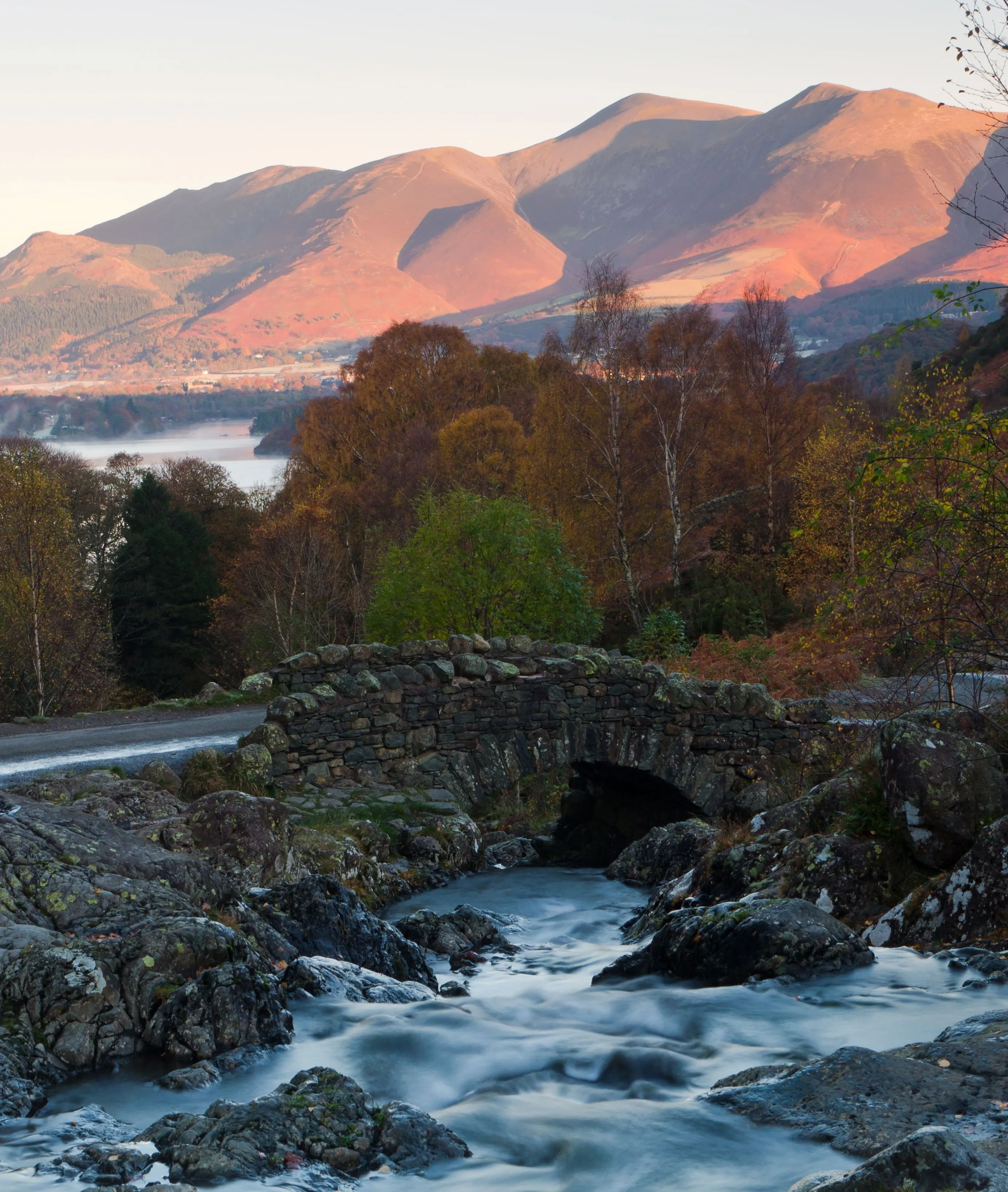 Stone bridge over a fast-flowing stream beneath sunlit mountains and autumn woodland in the Lake District.