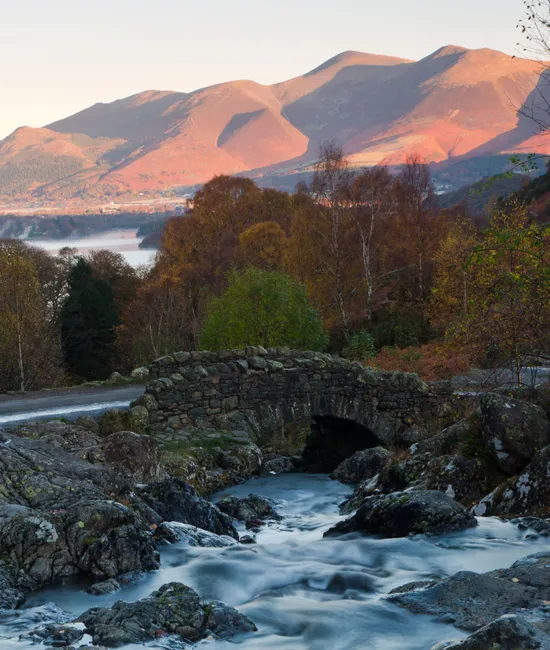 Stone bridge over a fast-flowing stream beneath sunlit mountains and autumn woodland in the Lake District.