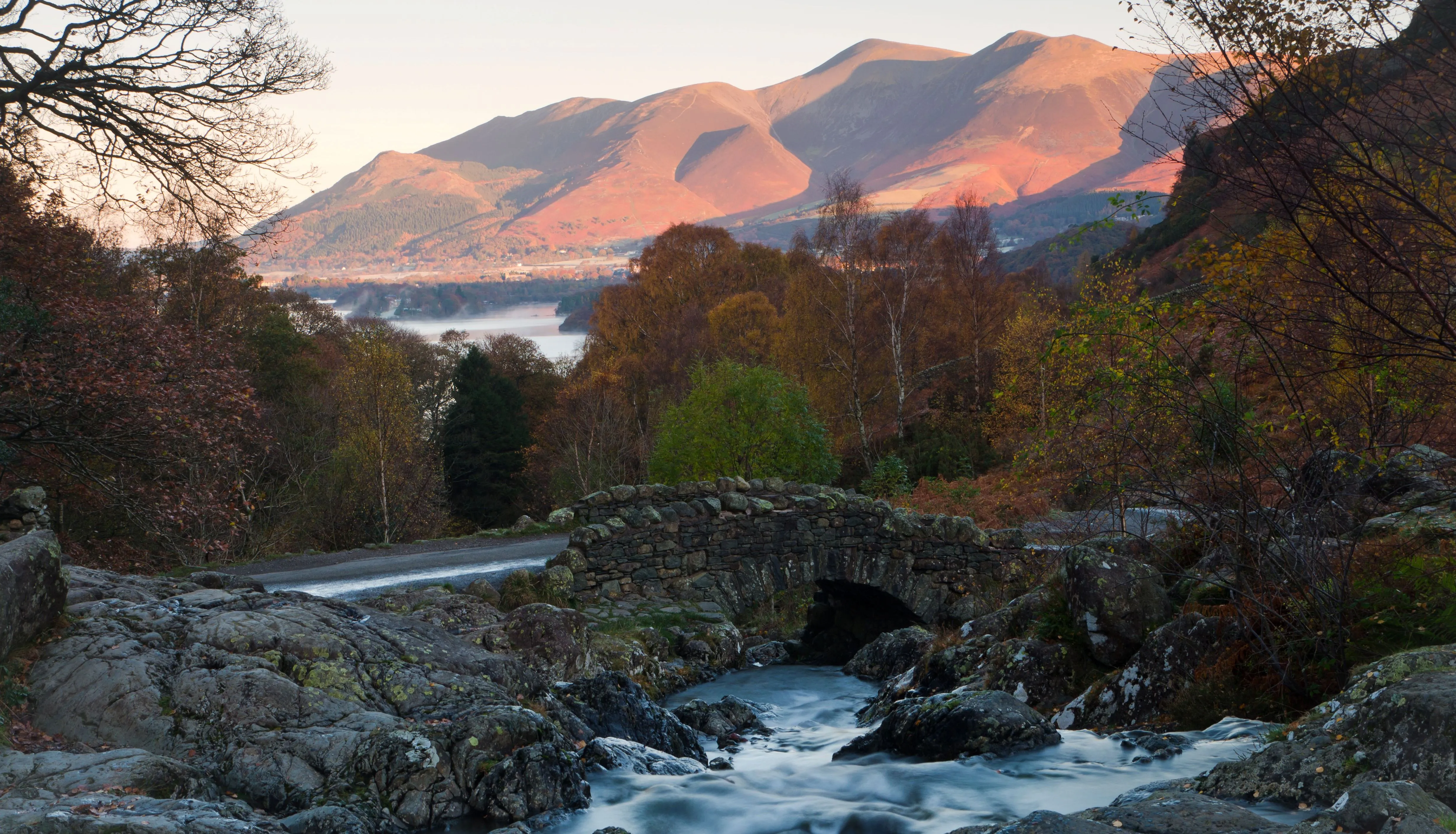 Stone bridge over a fast-flowing stream beneath sunlit mountains and autumn woodland in the Lake District.